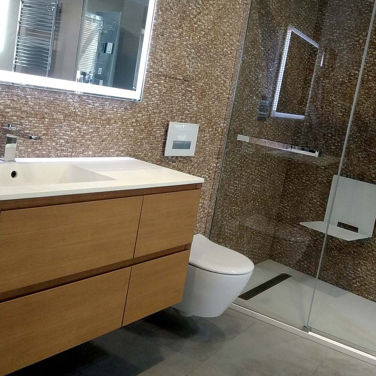 Bany amb pica i moble integrat de fust, WC i plat de dutxa gran amb mampara i rajoletes petites a tot el bany de colors marronosos.